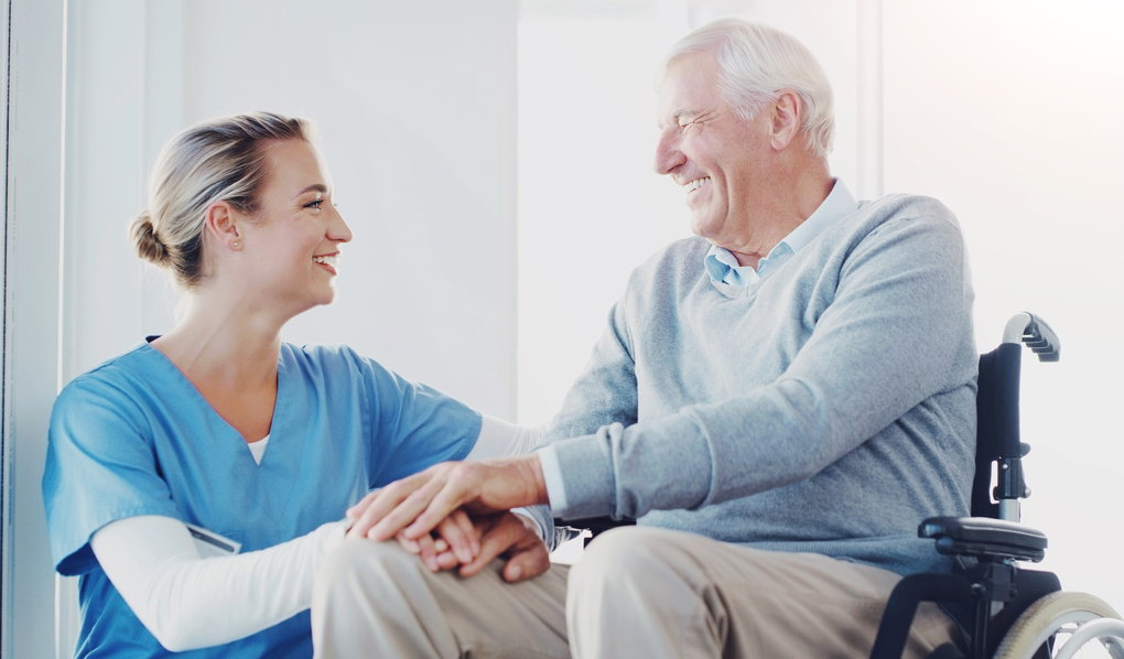 Caregiver, smile and senior patient in hospital, holding hands and compassion for old man with disability. Clinic, nurse and elderly person on wheelchair, care and support for healthcare and wellness